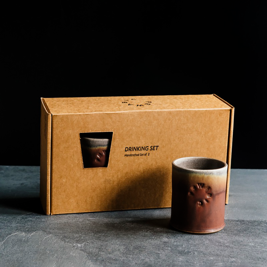 Box set of two toasty red-brown, Cinnamon Rocks Cups. The box reads "Drinking Set - Handcrafted Set of 2." One of the cups is outside of the box to showcase its glaze surface against a black backdrop and slate foreground. 