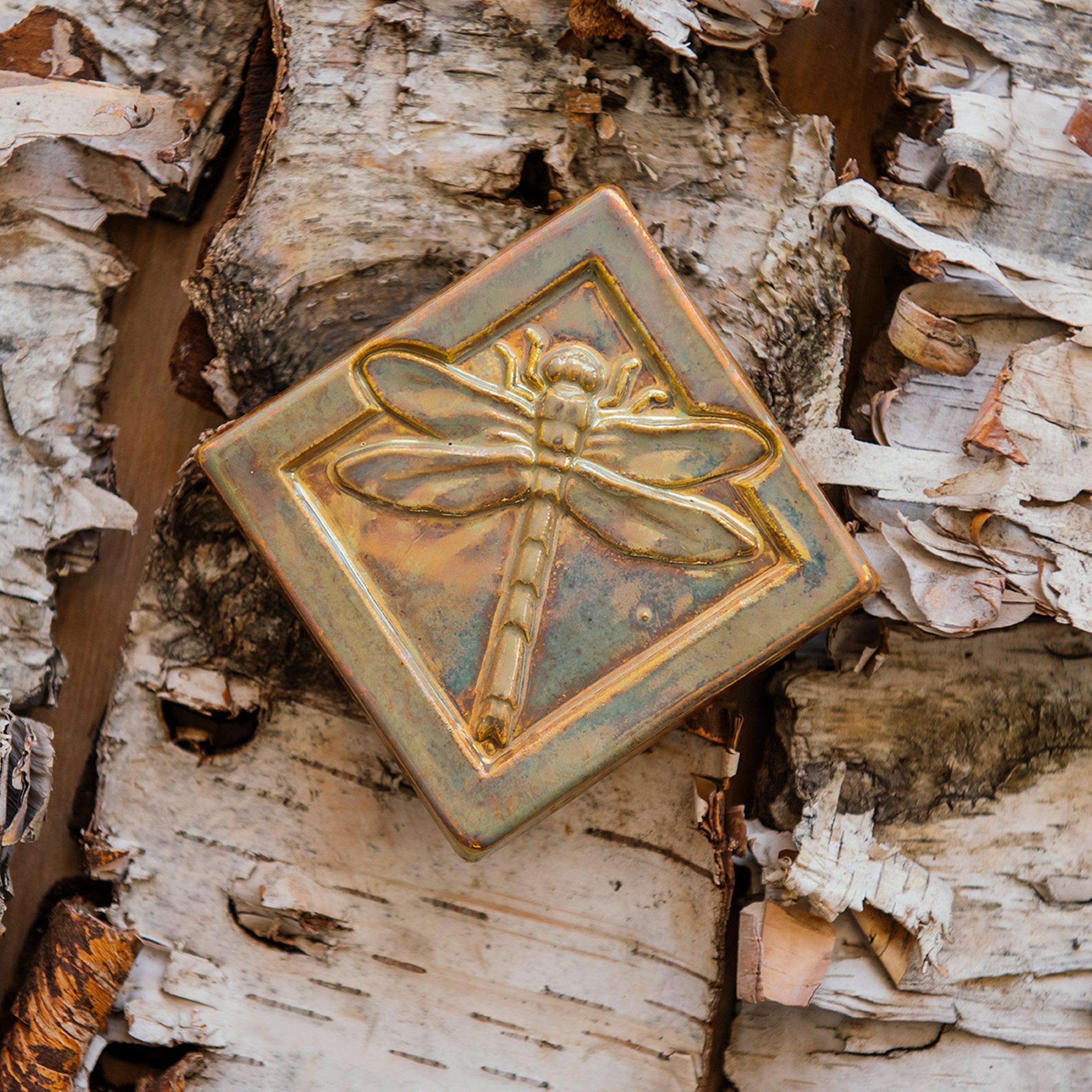 The Dragonfly Tile features a detailed dragonfly with wings outstretched. It is sitting horizontally on the square tile with a thick border around the edge. The pinkish gold metallic Blush Iridescent glaze features lots many variations. Depending on the lighting in the room, these pieces will pick up different hues.