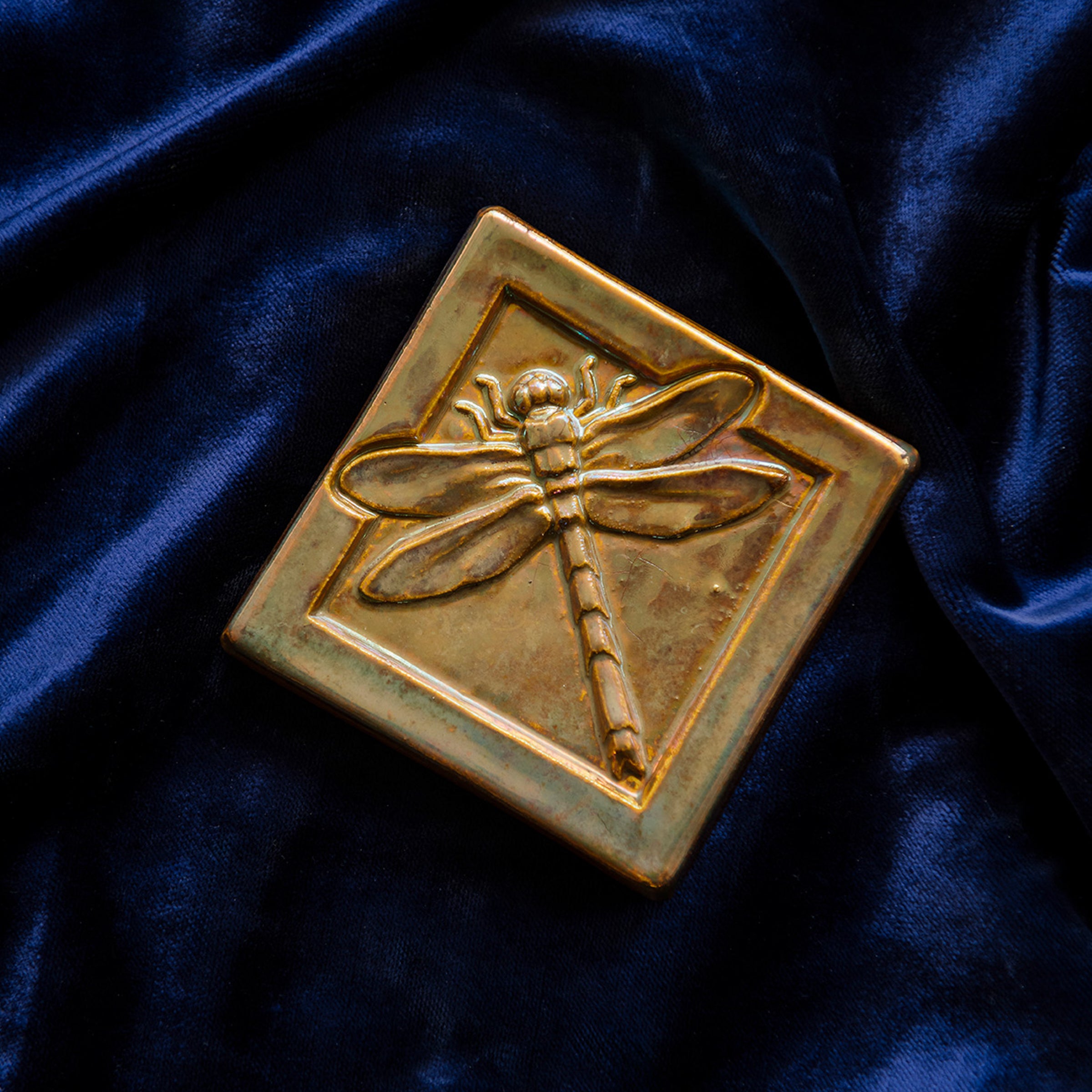 The Dragonfly Tile features a detailed dragonfly with wings outstretched. It is sitting horizontally on the square tile with a thick border around the edge. The pinkish gold metallic Blush Iridescent glaze features lots many variations. Depending on the lighting in the room, these pieces will pick up different hues.