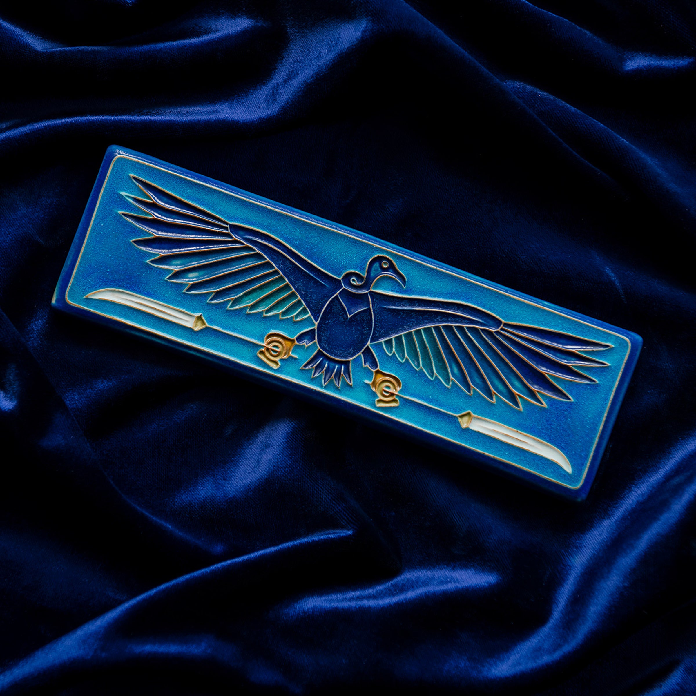 The short and wide Nekhbet tile features a line drawing of a vulture with wings outstretched, head turned to the right as she holds two stylized feathers- one in each talon. This tile features bright shades of blue. This tile rests on a bed of rich, blue velvet. 