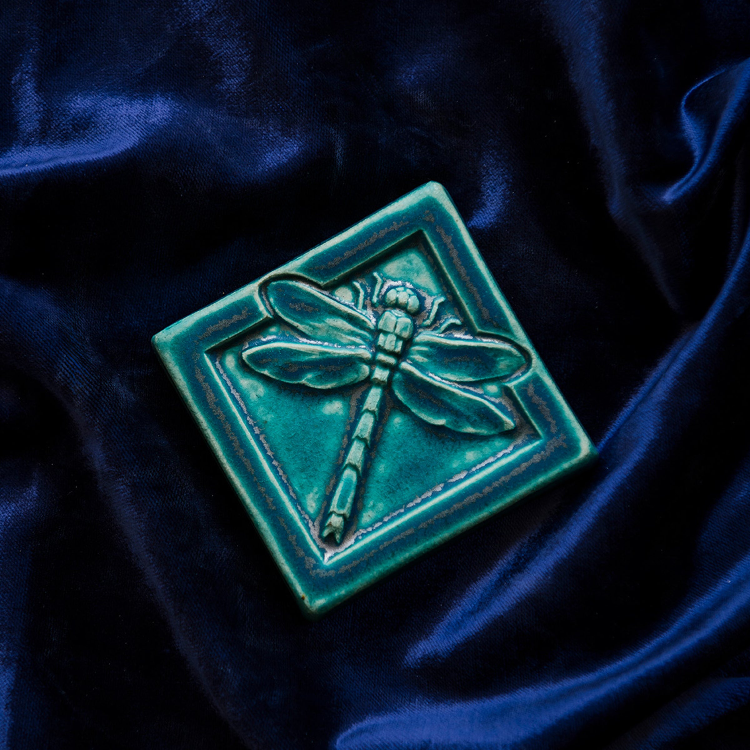 The Dragonfly Tile features a detailed dragonfly with wings outstretched. It is sitting horizontally on the square tile with a thick border around the edge. This Dragonfly Tile features the blueish-teal Pewabic Blue glaze.