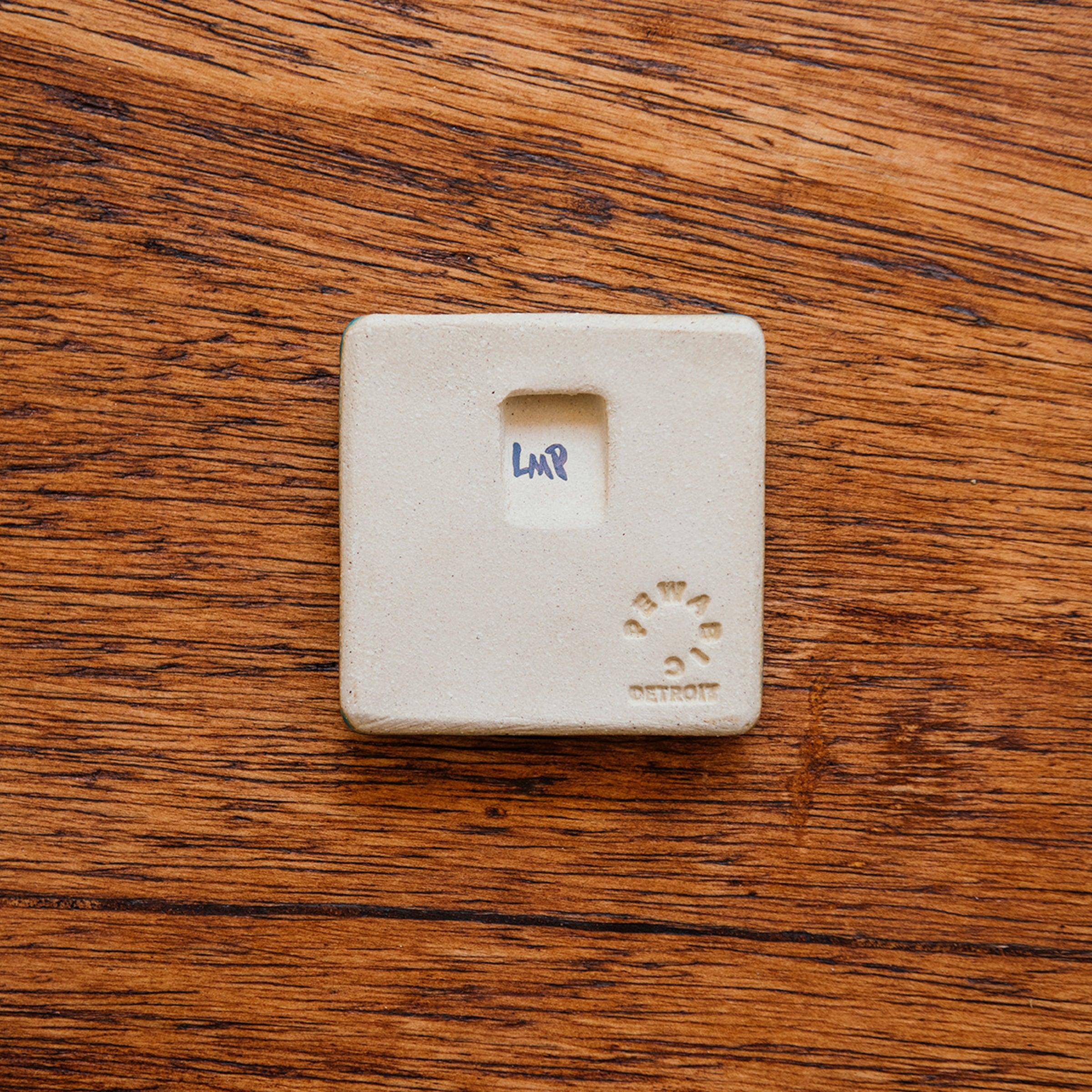 Back of a 3" x 3" tile with stamped-in notch for hanging. It is also stamped with the Pewabic logo, "Detroit", and specific Pewabic maker's mark. 