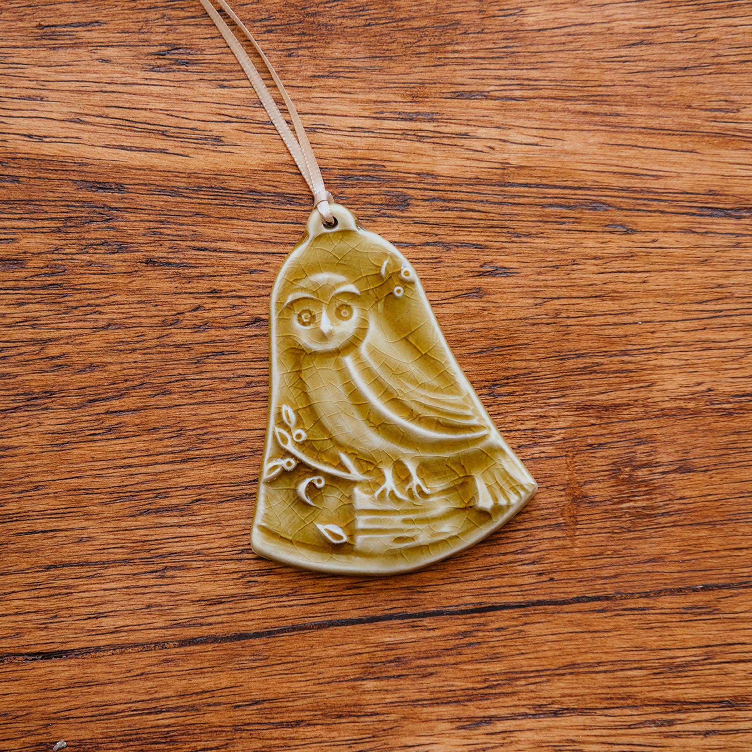 This bell-shaped ornament features a wide-eyed owl sitting on a fallen tree trunk, branches covered in leaves and berries surround it.