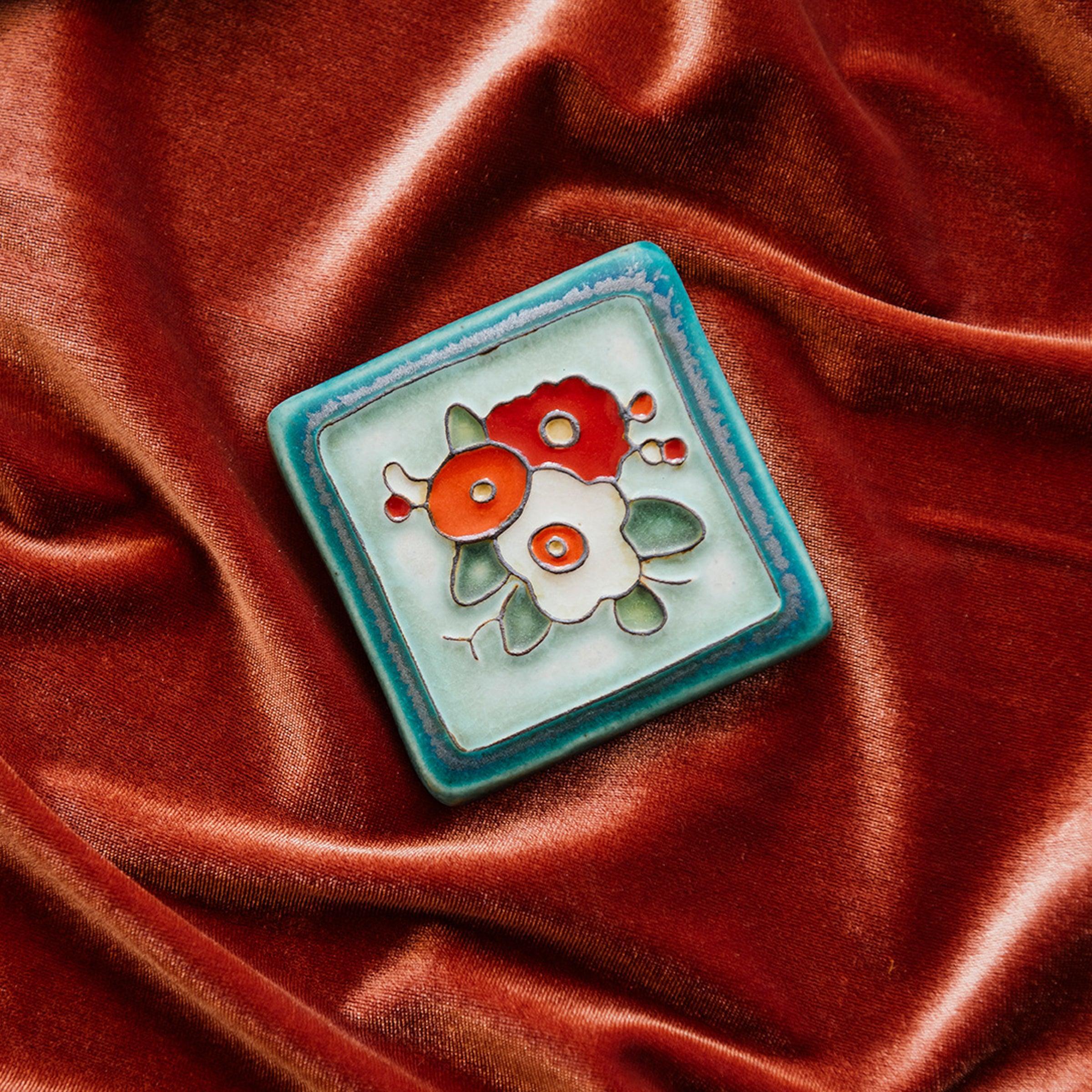 A Hand-Painted Three Flowers Tile in our "Poppy Palette" rests on a bed of rich, organge-pink velvet. 