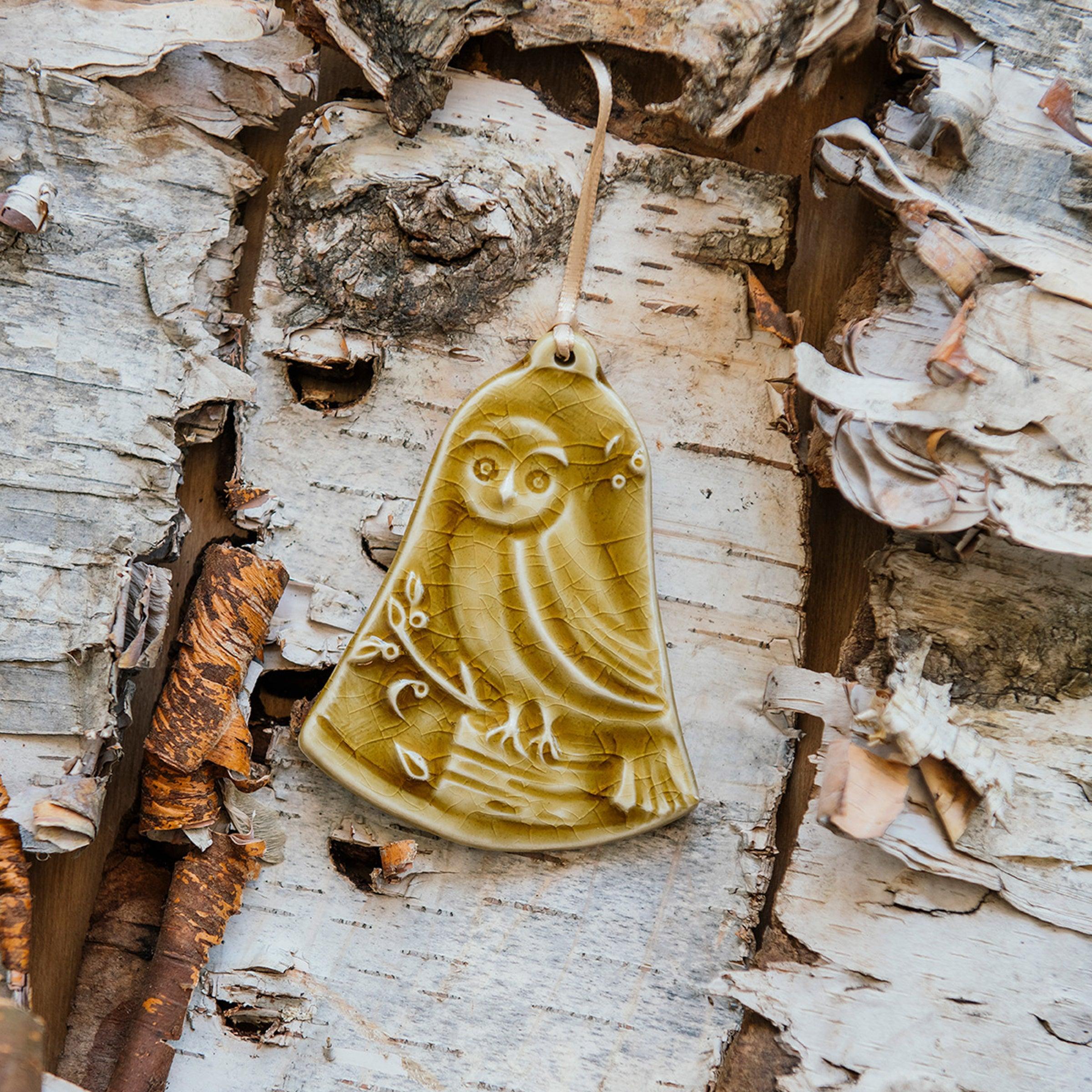 This bell-shaped ornament features a wide-eyed owl sitting on a fallen tree trunk, branches covered in leaves and berries surround it.