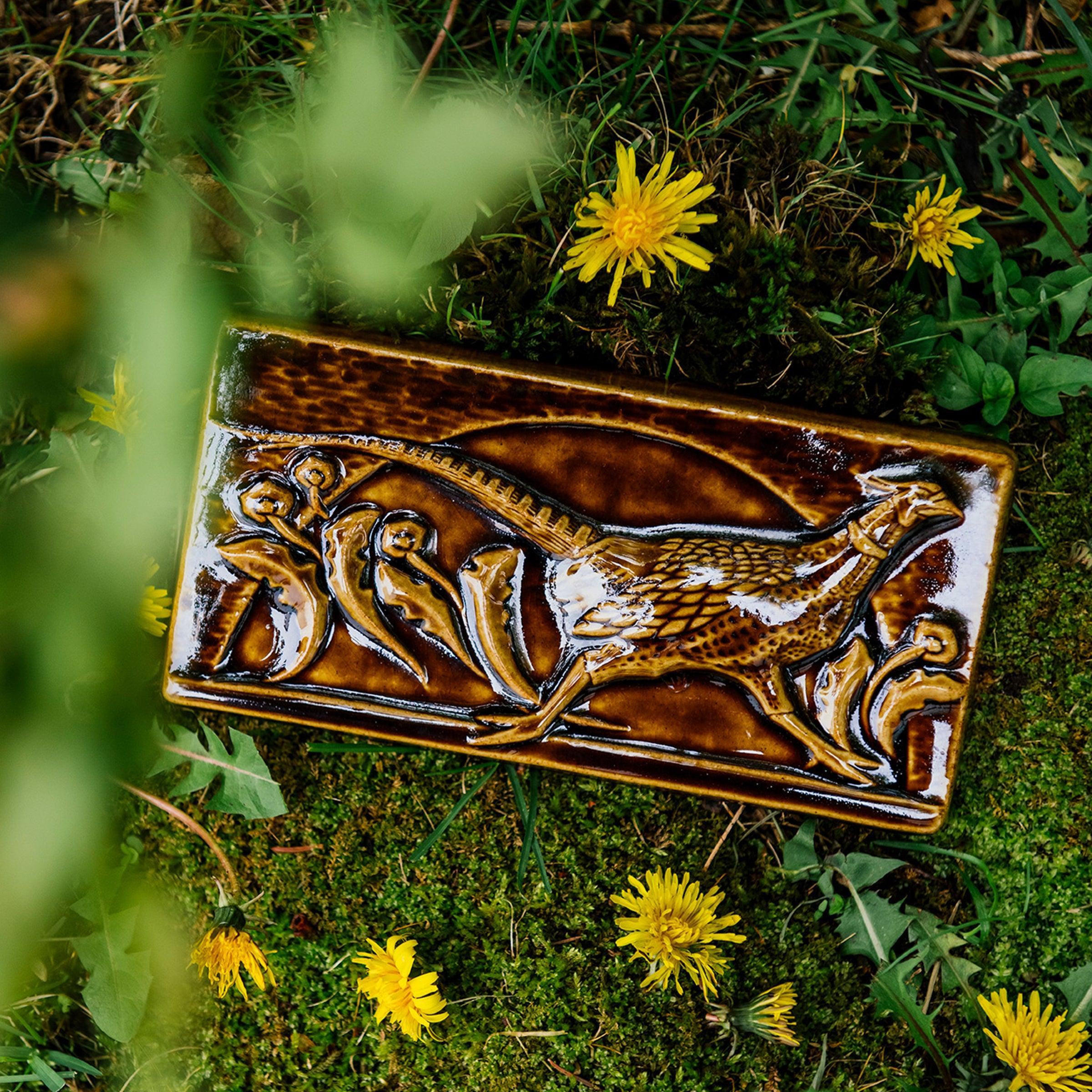 The long, horizontal Pheasant Tile features a male ring-necked pheasant running across the tiles. Puffy late-summer dandelions surround its outstretched feet. A large, domed sun is setting behind it. Featured in in a glossy brown Molasses glaze. 