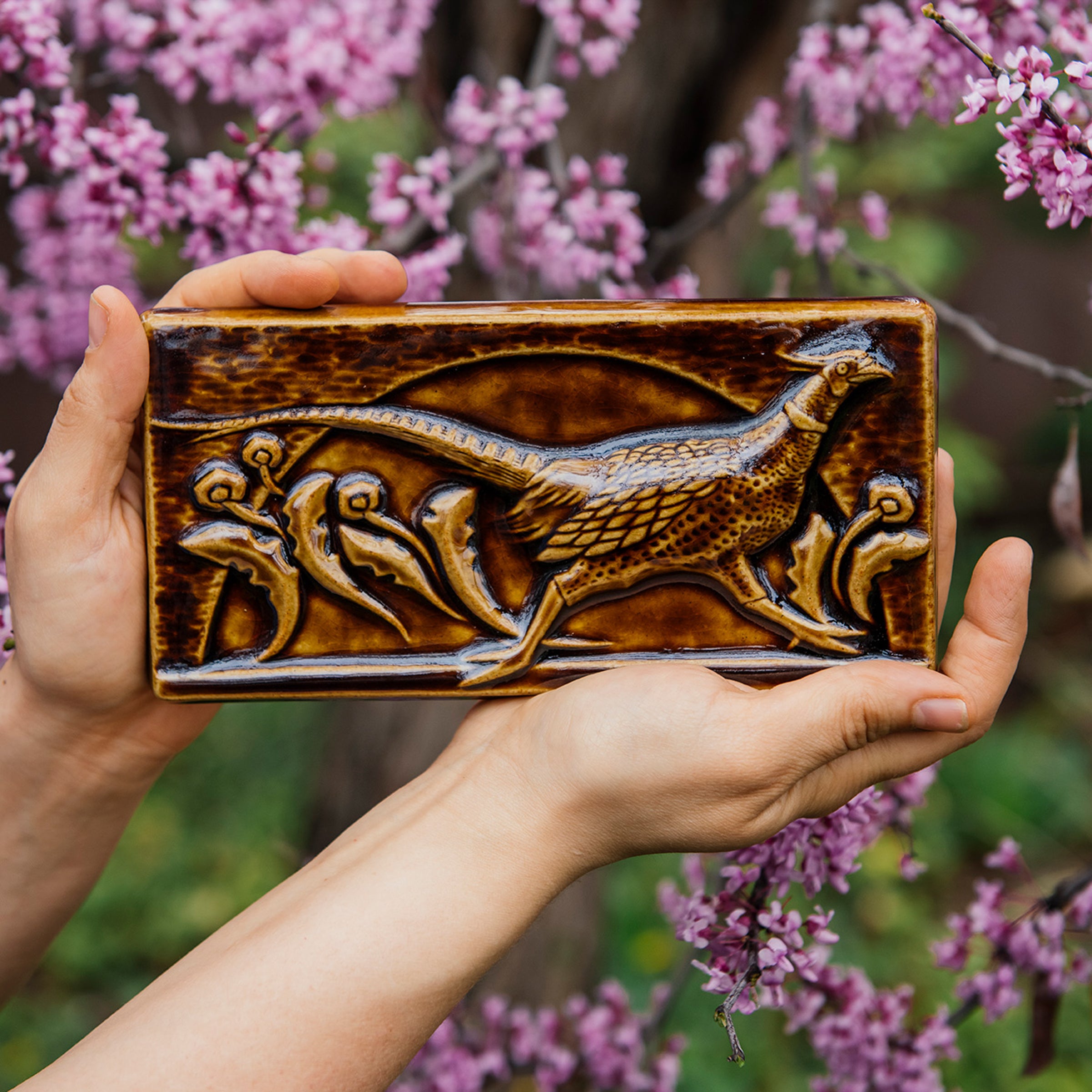 The long, horizontal Pheasant Tile features a male ring-necked pheasant running across the tiles. Puffy late-summer dandelions surround its outstretched feet. A large, domed sun is setting behind it. Featured in in a glossy brown Molasses glaze. 