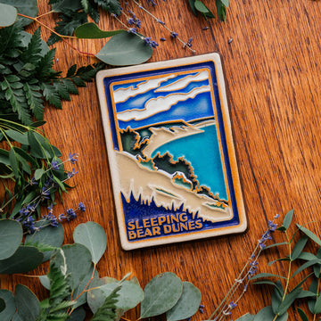 The Sleeping Bear Dunes Tile features the serene Lake Michigan and the sandy beaches and dunes covered on top by bushes and greenery. The words "Sleeping Bear Dunes" line the bottom of the design. The hand painted tile boasts blues and greens and pale yellow sand.