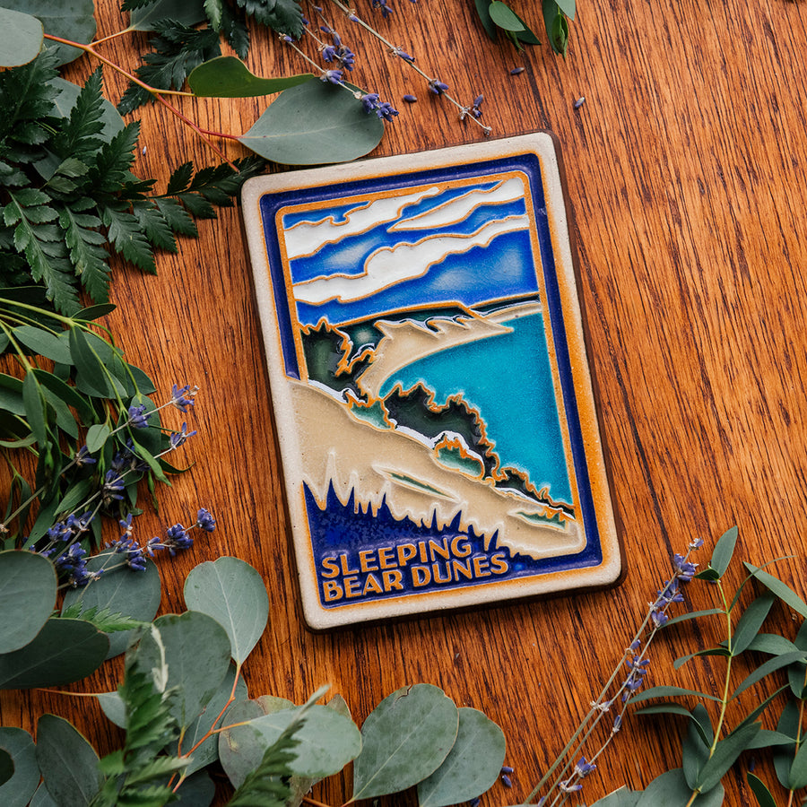 The Sleeping Bear Dunes Tile features the serene Lake Michigan and the sandy beaches and dunes covered on top by bushes and greenery. The words "Sleeping Bear Dunes" line the bottom of the design. The hand painted tile boasts blues and greens and pale yellow sand.