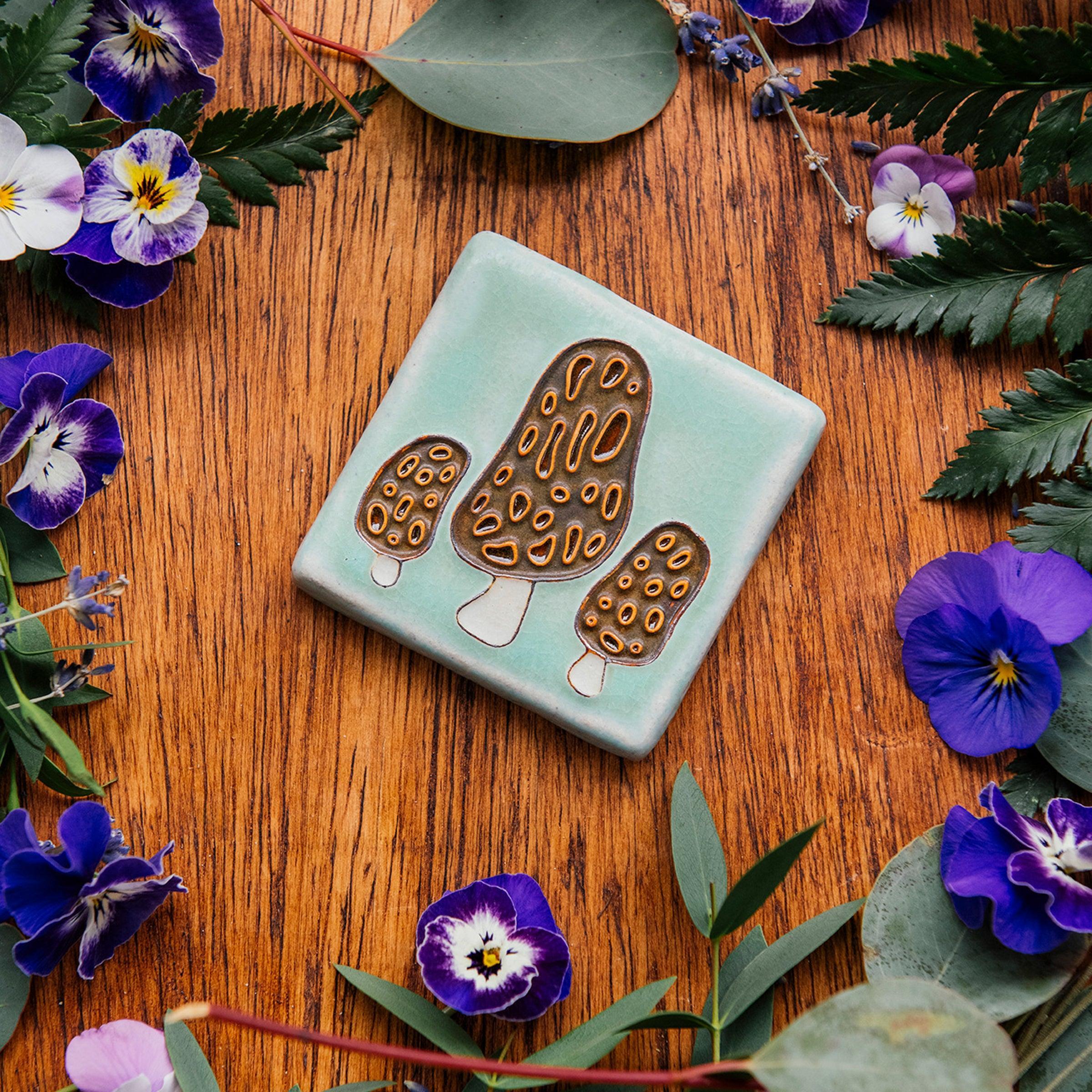 A tile depicting a line-drawn trio of brown Morel mushrooms on a light sea foam-glazed background. 