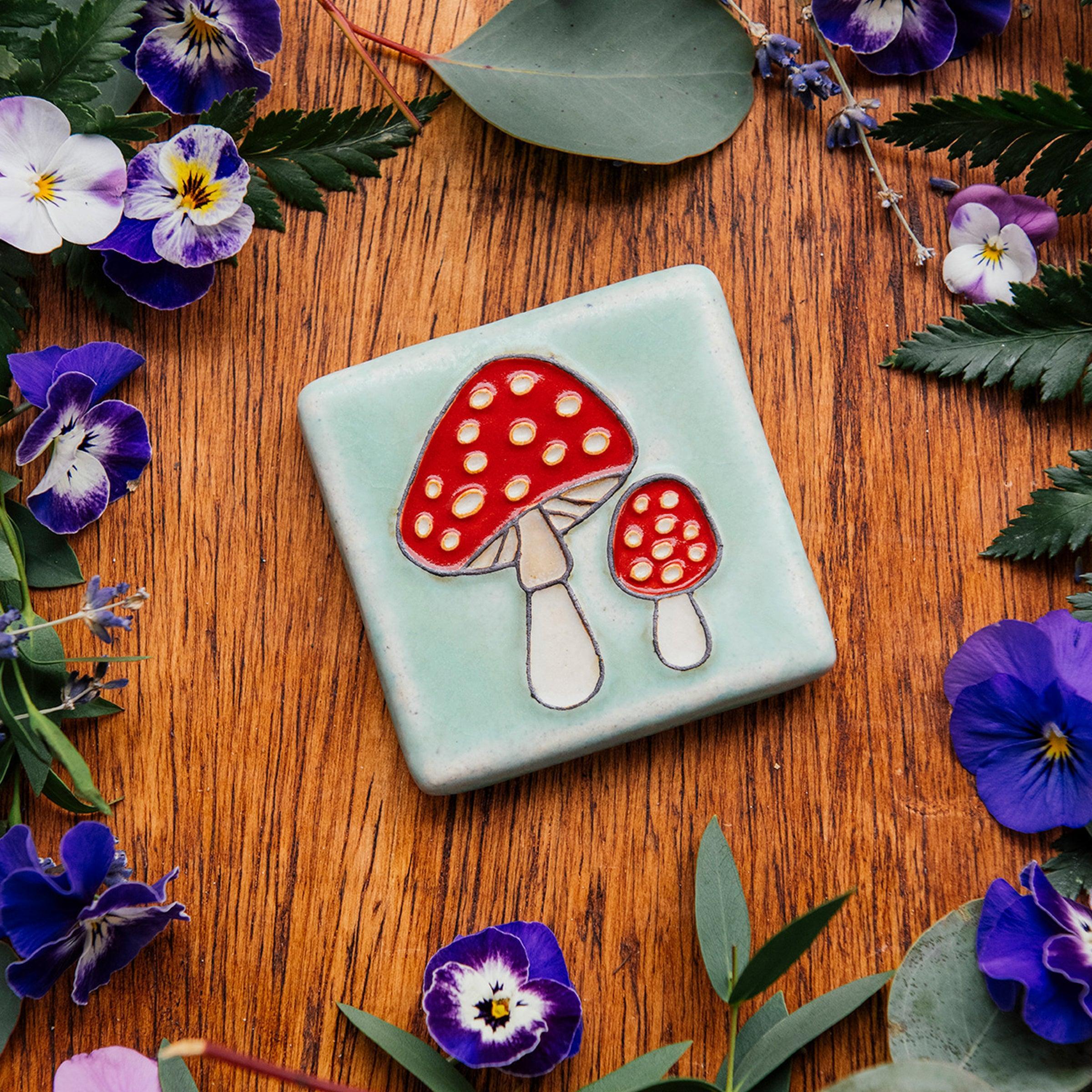 A hand-painted tile depicting a line drawing of a pair of Amanita mushrooms with classic red tops and white spots set on a light sea foam colored background glaze. 