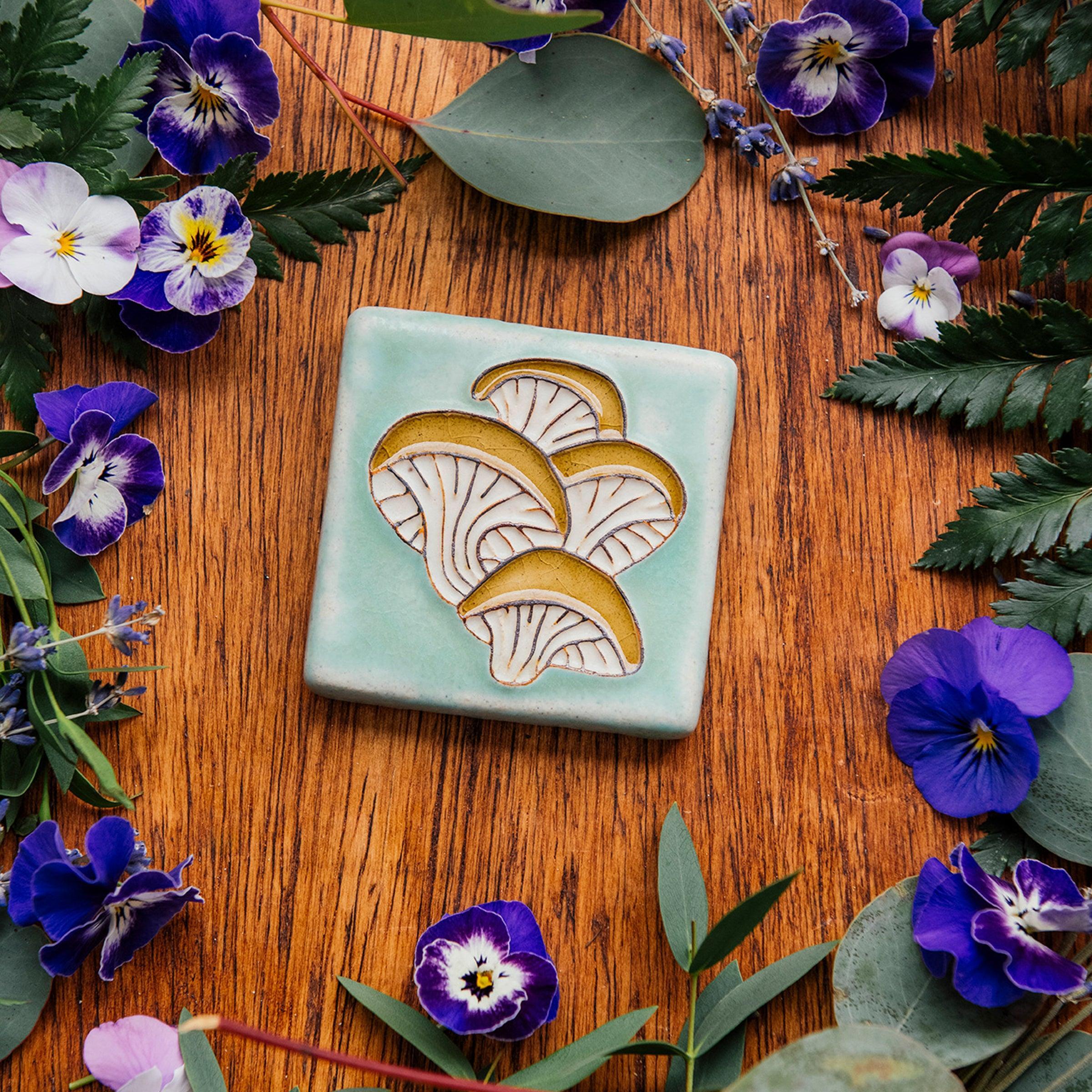 A hand-painted tile depicting a line drawing of Oyster mushrooms with golden yellow tops and white bases set on a light sea foam colored background glaze. 