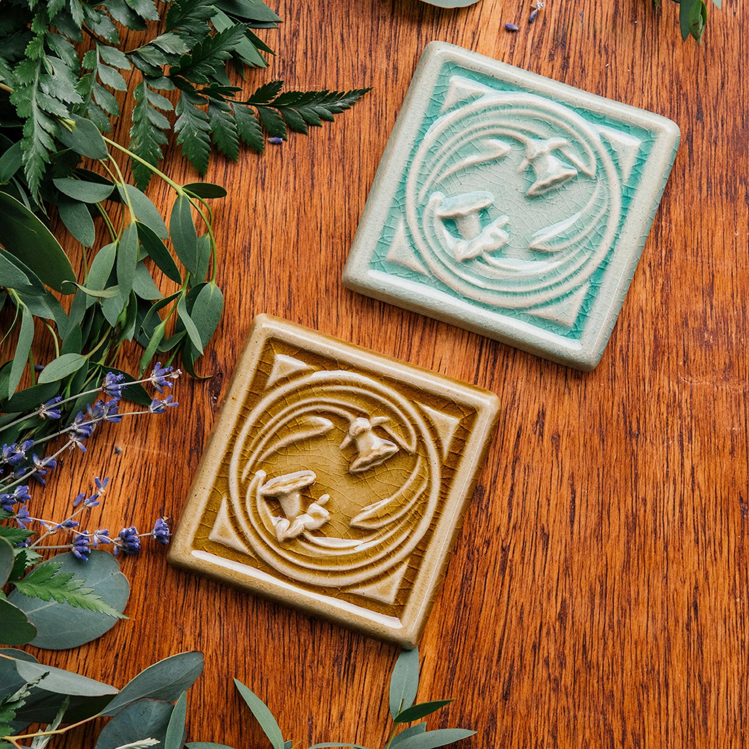A pair of Daffodil Tiles rest on a beautiful wood finish surrounded by summer greenery. They are glazed in our glossy Celadon and Honey glaze. 