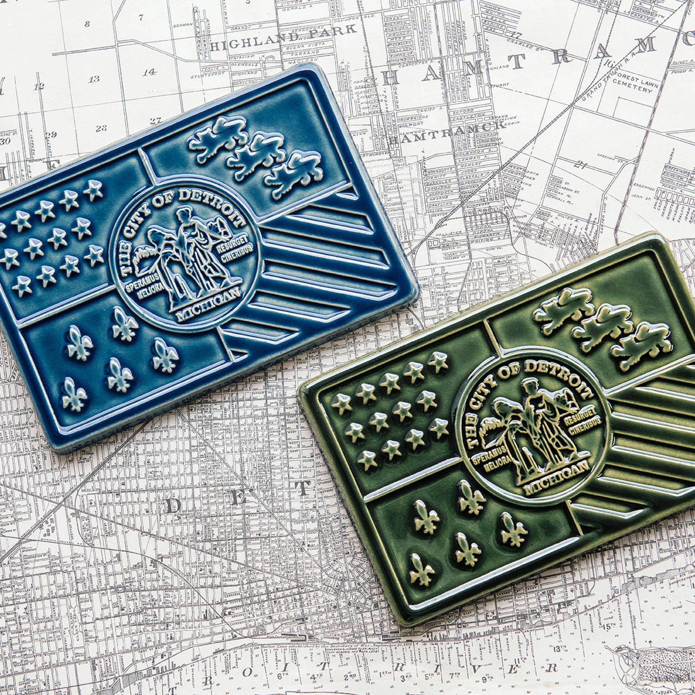 The Detroit Flag Tile is a ceramic version of the city of Detroit's flag. It is divided into four rectangular sections, one in each corner, that feature stars, fluer-de-lis, stripes and lions. In the center sits a round seal featuring a detailed design. 