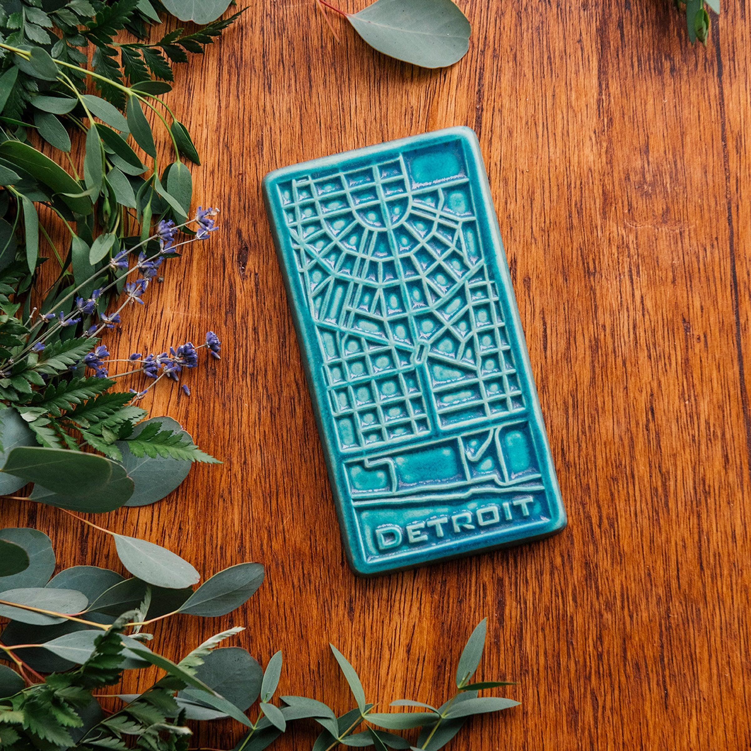 This Pewabic Blue Detroit Map Tile is sitting on a wooden board, surrounded by flowers and eucalyptus. 