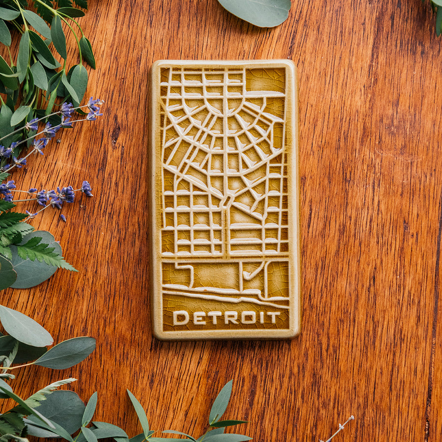 This Honey Glazed Detroit Map Tile is sitting on a wooden board, surrounded by flowers and eucalyptus. 