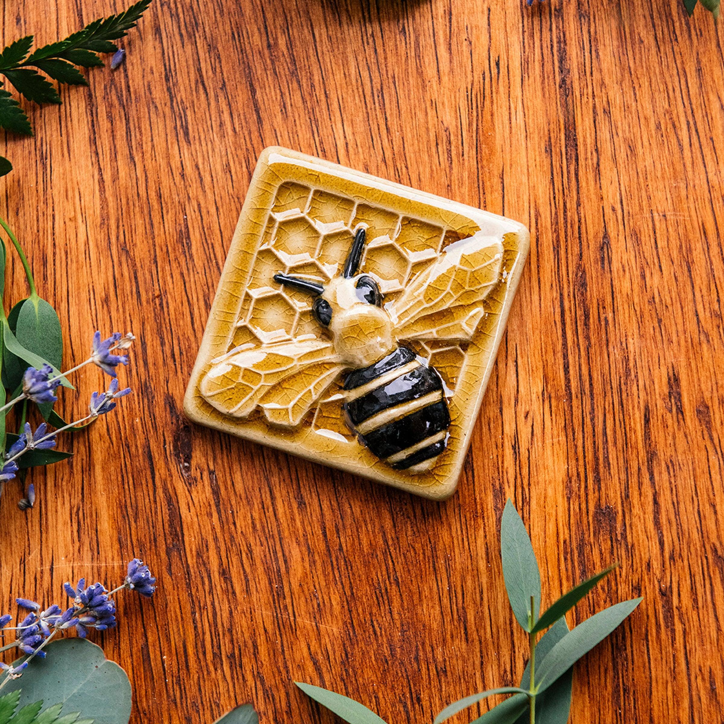 A glossy hand-painted Honeybee Tile in golden honey and black details surrounded lavender and summer greenery. 