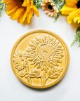 The Sunflower Trivet Tile features two sunflowers growing up into the circular tile with leaves and a bit of stalk in view. One sunflower is facing forward while the other is slightly smaller and facing to the left.