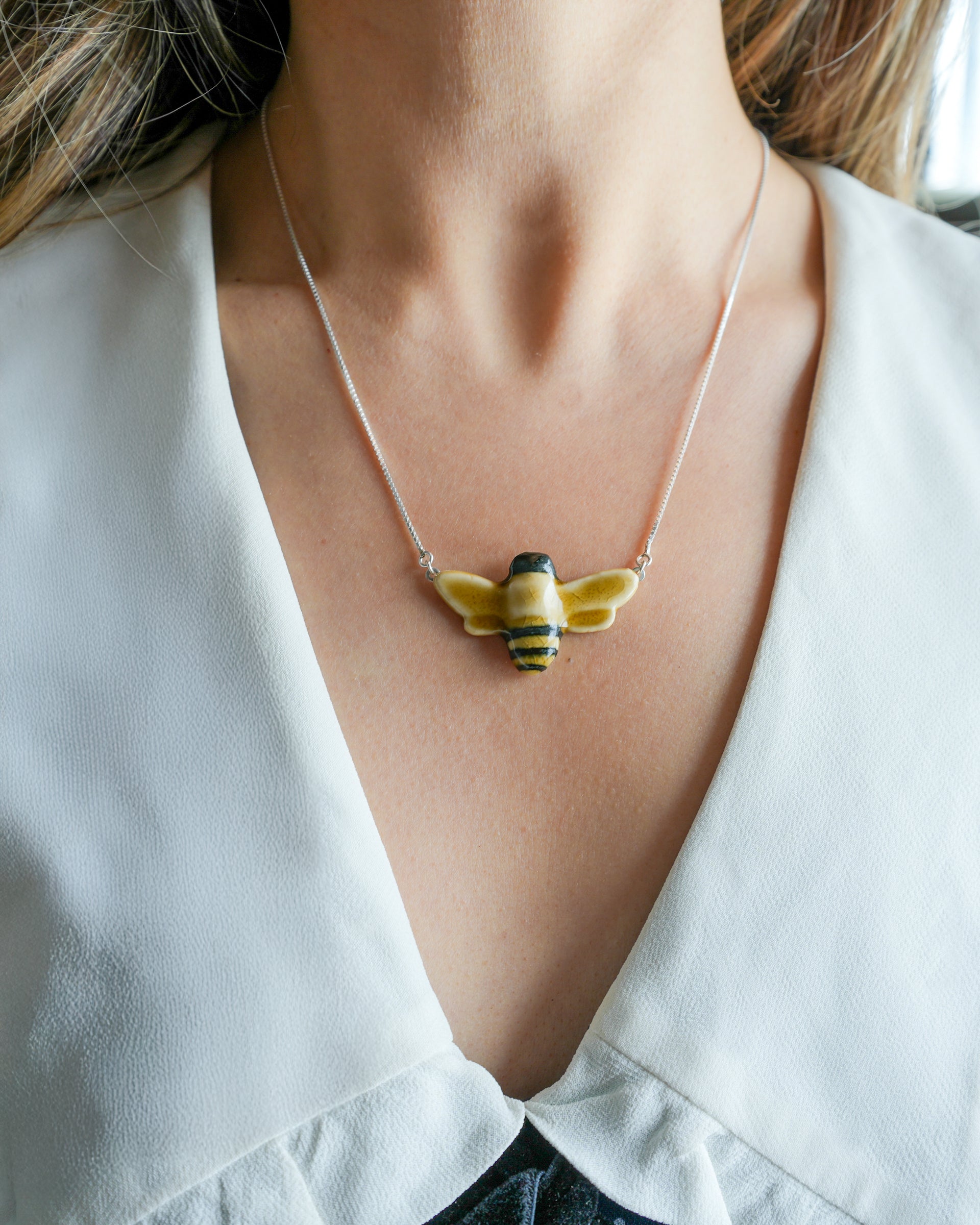 Necklace with a glossy, hand-painted bee pendant worn by a person wearing a dress with a large, white collar. 