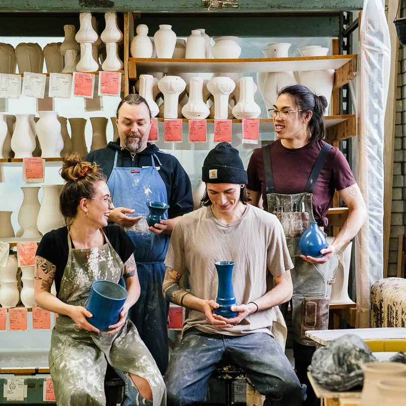 Pewabic's vessel making team poses in front of a shelving unit full of their thrown vessels and slip cast vases. They look at each other and smile, each one holding piece that they created.