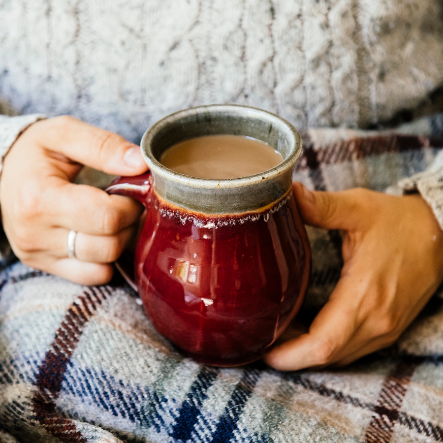 The smooth exterior of the mug shines in the sunlight while a woman in a warm sweater grasps it in her hands.