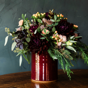The crock is a cylindrical vessel with straight sides that has a wide opening and is as wide as it is tall. This crock is filled with a large bouquet of burgandy and white flowers.