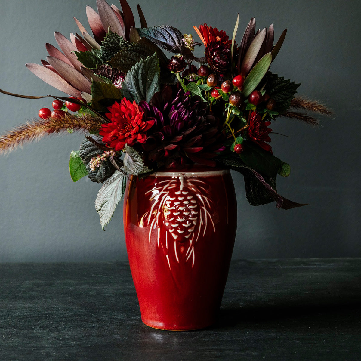 The Pinecone vase features smooth sides with three high relief pinecones and pine needles near the lip of the vessel. Each pinecone points in a different direction so there is always one pinecone pointed outword no matter which direction the vase is sitting. The top of the vase has a simple raised lip that come in toward the center.