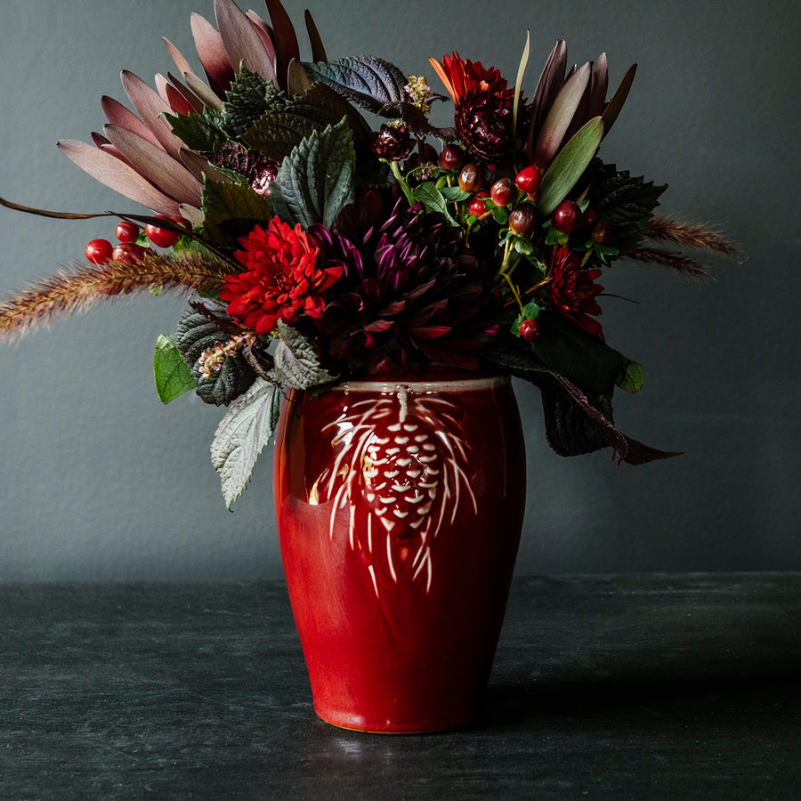 The Pinecone vase features smooth sides with three high relief pinecones and pine needles near the lip of the vessel. Each pinecone points in a different direction so there is always one pinecone pointed outword no matter which direction the vase is sitting. The top of the vase has a simple raised lip that come in toward the center.