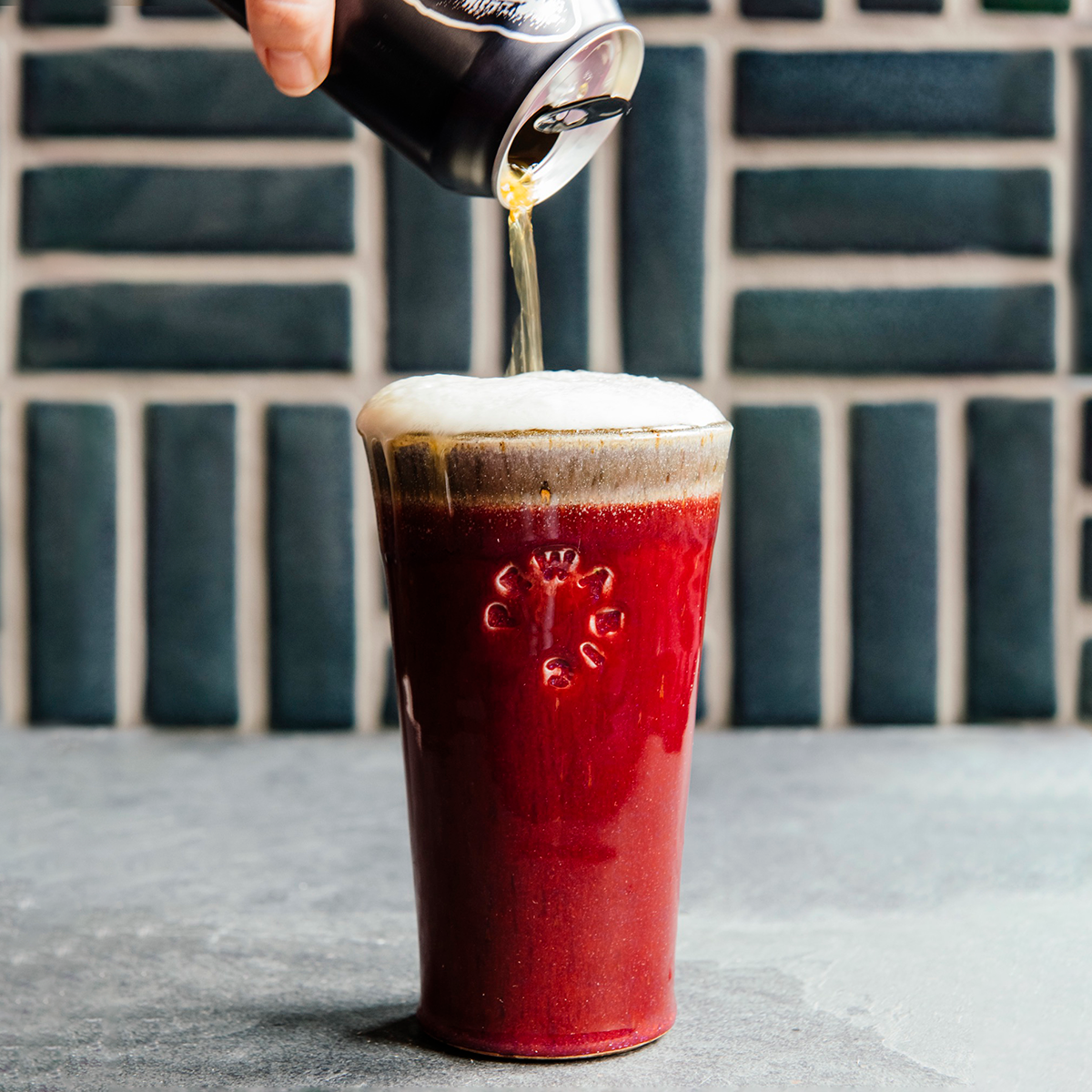 The Pewabic Pint is a simple ceramic pint glass with sides that slightly slope outwards to create a larger opening at the top. These pints feature smooth sides and have been stamped with the circular Pewabic logo on one side. The the glossy, deep red Winterberry glaze is featured on the sides of the cup with a warm gray glaze inside that spills over about an inch onto the outer cup, with the two colored glazes meeting just above the logo.