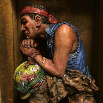 This realistic ceramic sculpture shows a man wearing a denim cut-off vest and coveralls open to the waist. He sits with his chin resting on his clasped hands which are stacked on a sticker-covered worker's helmet.