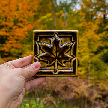 A person's hand is reaching into the bottom-left of the frame holding a Maple Leaf tile in a deep, glossy Molasses glaze. There are trees in the background with vibrant, autumn leaves. 