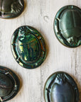 Five paperweights rest in a haphazard fashion - they feature the Matte Green Iridescent glaze. Each tile exemplifies the variations found in this glaze. Some are a shiny metallic while others are more matte.