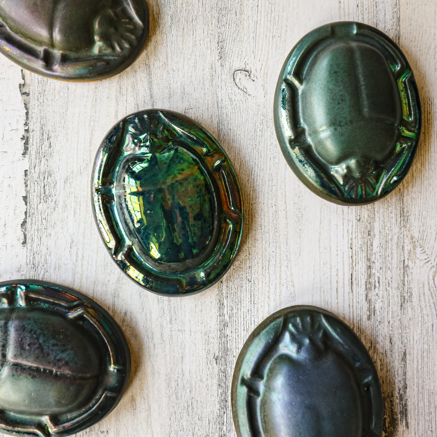 Five paperweights rest in a haphazard fashion - they feature the Matte Green Iridescent glaze. Each tile exemplifies the variations found in this glaze. Some are a shiny metallic while others are more matte.