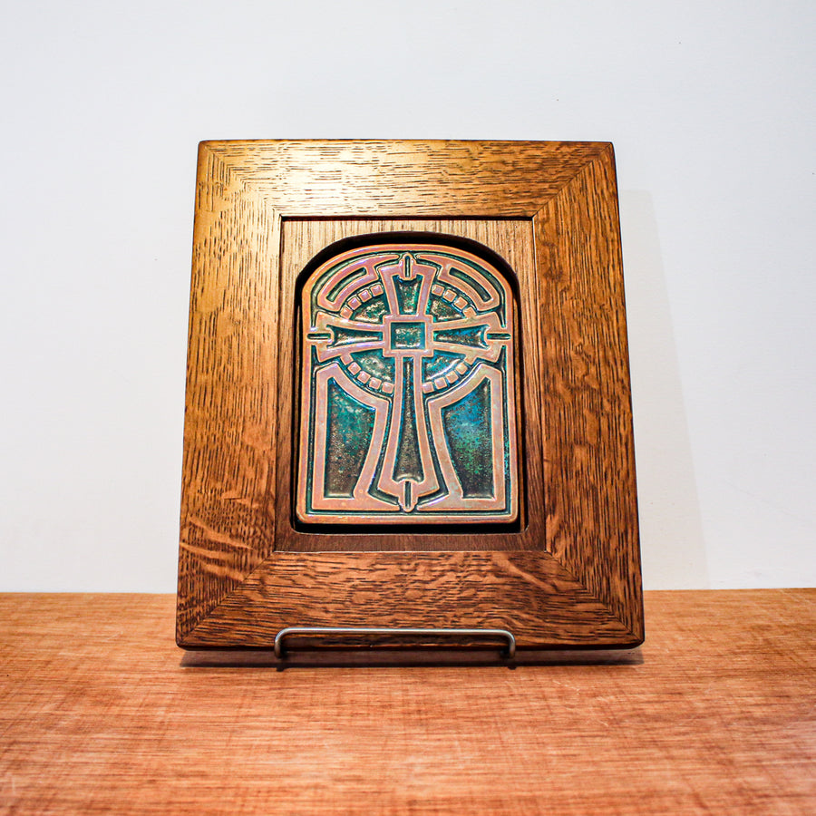 The Cross Tile has rounded edges at the top, creating a look similar to a stained glass window. The cross itself is ornate with a halo behind it. The cross and ornate details features the gold Blush Iridescent glaze while the background of the tile is in the matte, brushed metal green of the Matte Green Iridescent glaze. The frame hugs the tile at the top, the wood rounding to frame the curved nature of the tile. The oak frame is in a yellowish brown tone.