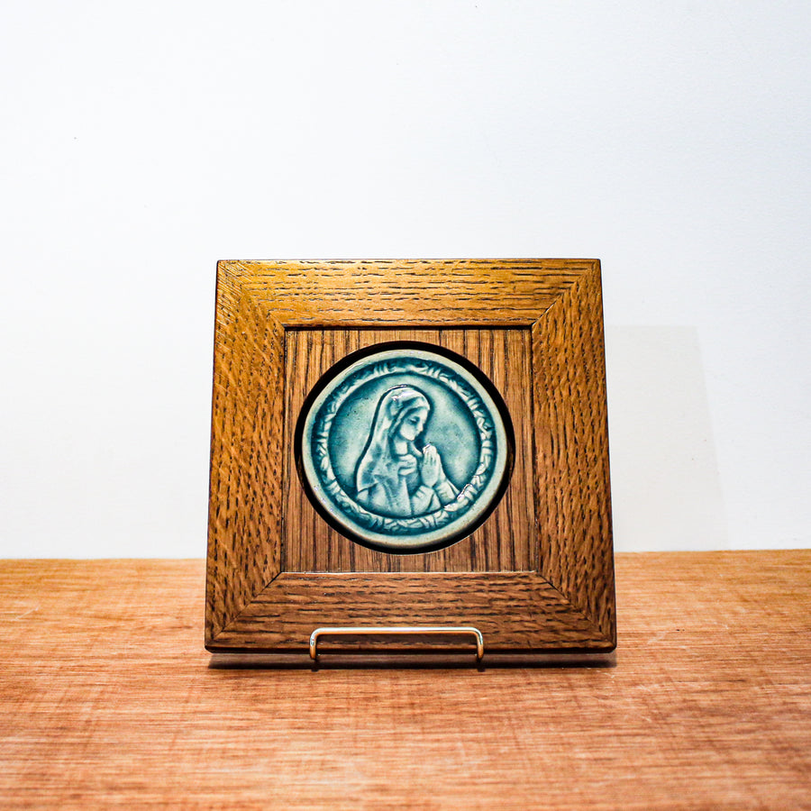 This circular ceramic Blessed Virgin Mary tile depicts Mary with head bowed and hands pressed together in prayer. The tile features the medium blue Glacier Gloss glaze which beautifully offsets the yellowish brown hue of the oak wood frame.