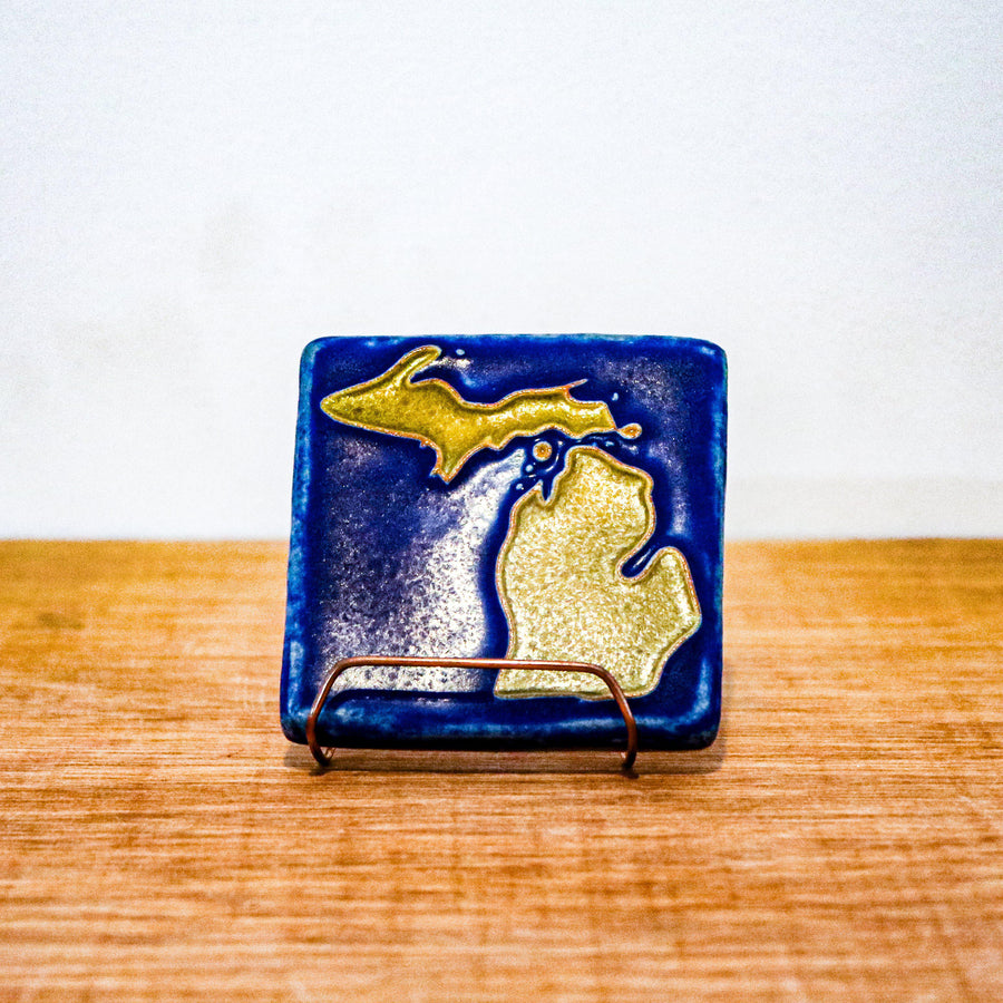 The Michigan Outline Tile features the outline of the state of Michigan with both peninsulas and its larger northern islands. This hand painted tile has a background of deep matte blue while the peninsulas are a maize gold color.