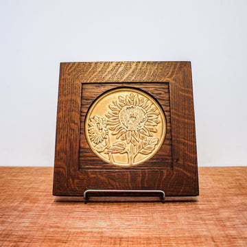 The Sunflower Trivet Tile features two sunflowers growing up into the circular tile with leaves and a bit of stalk in view. One sunflower is facing forward while the other is slightly smaller and facing to the left. This tile is in the matte golden yellow Mustard glaze which beautifully offsets the reddish brown oak wood frame.