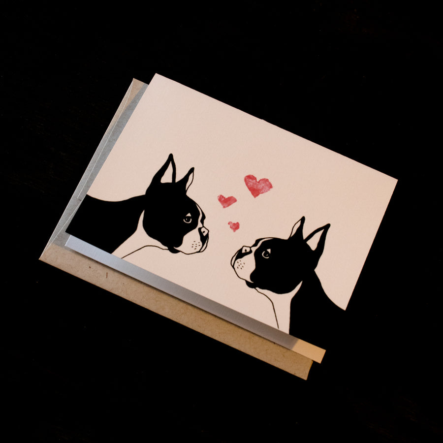 This white greeting card features two illustrated Boston Terriers staring at each other, there are three hearts floating above them.