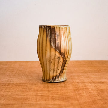 This Justin Lambert cup features a vase-like cup whose diameter starts smaller at the bottom, gets gradually wider then smaller again near the rim. There are vertical ridges and a moddled brown glaze.