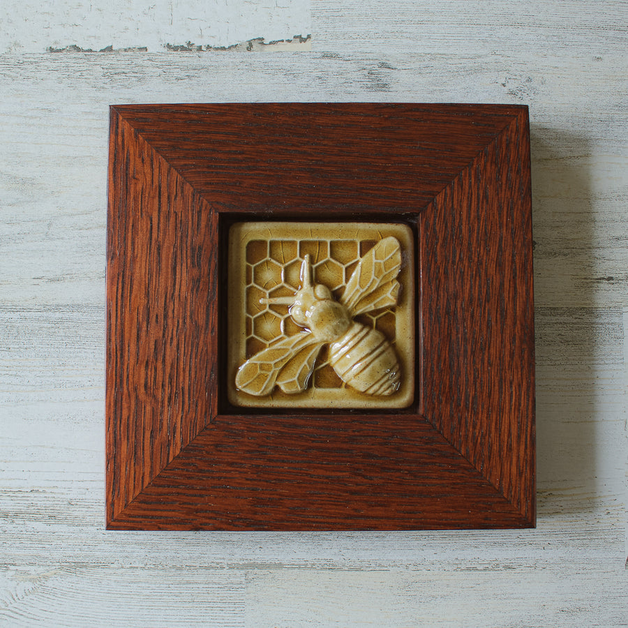 The Honeybee Tile features a large honeybee with stripes and detailed segmented wings. The bee sits on a honeycomb patterned background. The tile is in our deep golden Honey Gloss glaze which is beautifully offset by the deep reddish brown oak wood frame.