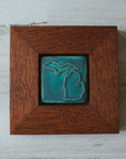The Michigan Outline Tile features the outline of the state of Michigan with both peninsulas and its larger northern islands. It is in the matte turquoise Pewabic Blue glaze which is beautifully offset by the reddish brown oak wood frame.