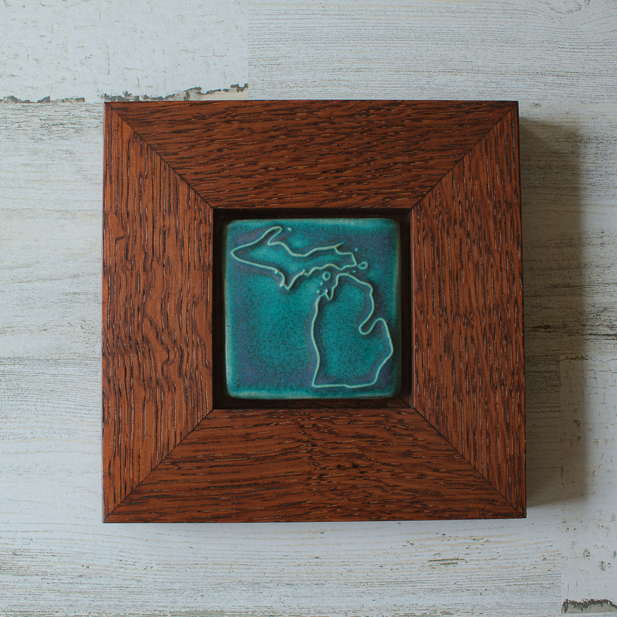 The Michigan Outline Tile features the outline of the state of Michigan with both peninsulas and its larger northern islands. It is in the matte turquoise Pewabic Blue glaze which is beautifully offset by the reddish brown oak wood frame.