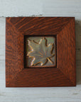 The Maple Leaf Tile features a simple maple leaf set into the square tile diagonally- with the stem and tip in opposite corners. The tile is in the pinkish gold metallic Blush Iridescent finish which has many variations. Depending on the lighting in the room, these pieces will pick up different hues. The oak frame is in a deep reddish brown color.