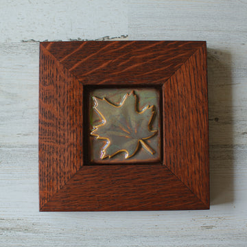 The Maple Leaf Tile features a simple maple leaf set into the square tile diagonally- with the stem and tip in opposite corners. The tile is in the pinkish gold metallic Blush Iridescent finish which has many variations. Depending on the lighting in the room, these pieces will pick up different hues. The oak frame is in a deep reddish brown color.