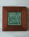 The Detroit Skyline tile features the current downtown skyline with the Detroit River in the foreground. A freighter floats by causing ripples on the otherwise flat surface of the water. The word "Detroit" is written above in the sky. This tile features the matte blueish-green Pewabic Green glaze which beautifully offsets the deep reddish brown oak frame.