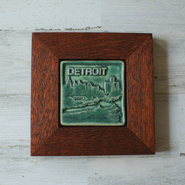 The Detroit Skyline tile features the current downtown skyline with the Detroit River in the foreground. A freighter floats by causing ripples on the otherwise flat surface of the water. The word "Detroit" is written above in the sky. This tile features the matte blueish-green Pewabic Green glaze which beautifully offsets the deep reddish brown oak frame.