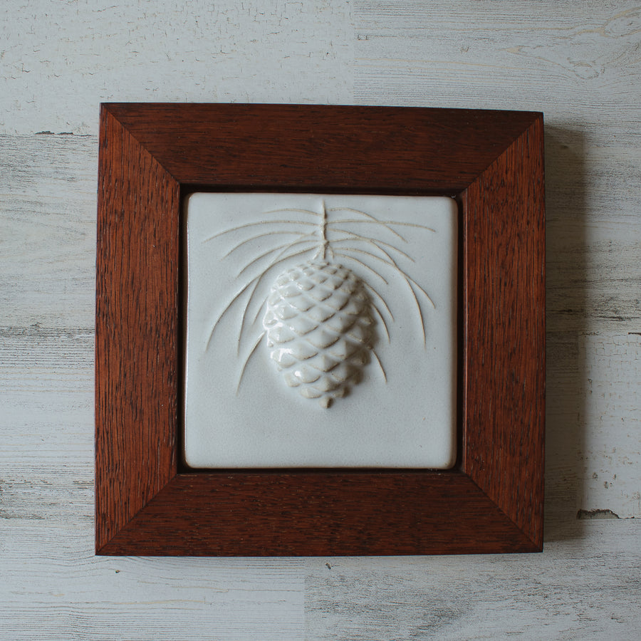 The ceramic Pinecone Tile has a high relief design with one large pinecone in the center and long spindly pine needles framing it. This tile is in the satin finished white Alabaster glaze which beautifully offsets the deep reddish brown hue of the oak frame.