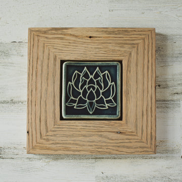 The Lotus tile features a line drawing of a blooming lotus flower, its pointed petals outstretching. It has a simple border around the design. A pale reclaimed wood frame holds the tile in the glossy deep blue Ocean glaze.