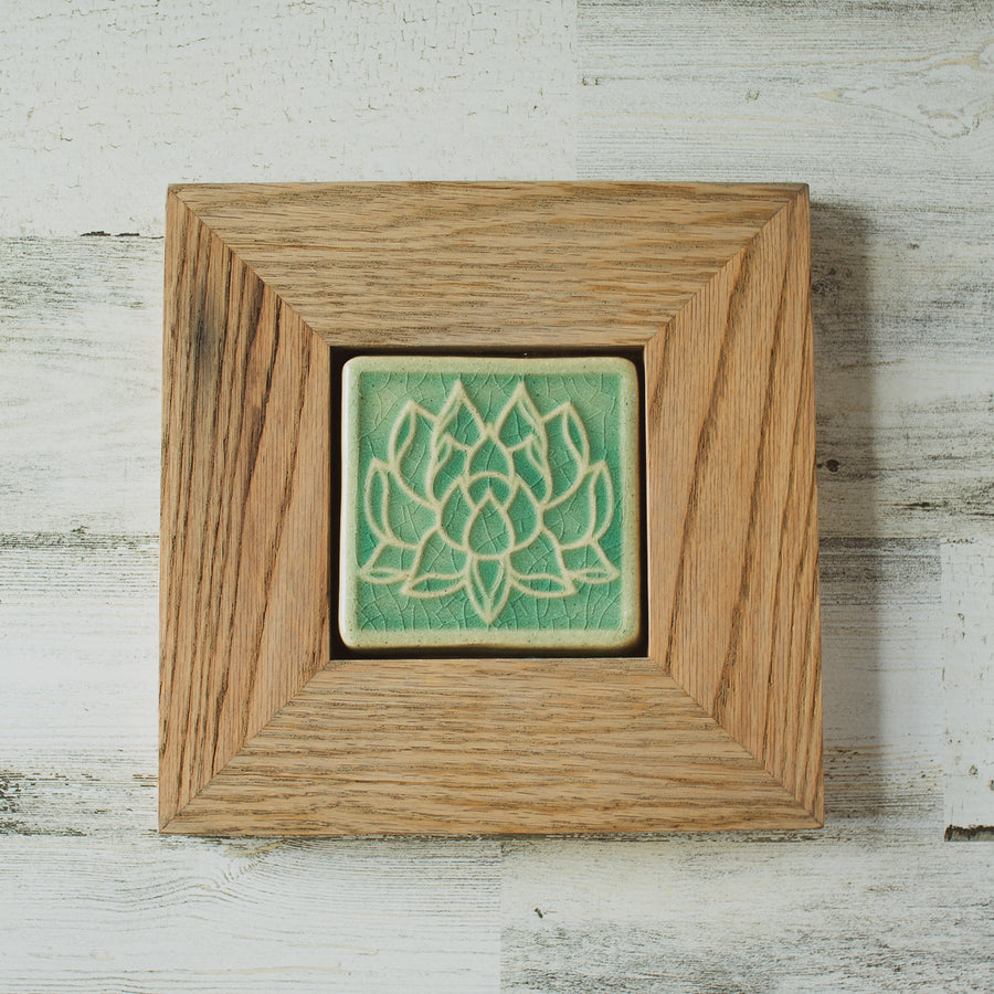 The Lotus tile features a line drawing of a blooming lotus flower, its pointed petals outstretching. It has a simple border around the design. A pale reclaimed wood frame holds the tile in the glossy pale blue Celadon glaze.