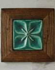 The Flower Geo Tile features a four-petaled flower. Each petal takes up a quarter of the tile, making a perfectly geometric shape. The inner part of the flower is round with four cattail shaped stamen exploding from the center that point out to the corners of the tile. This tile is framed in a cool colored wood.